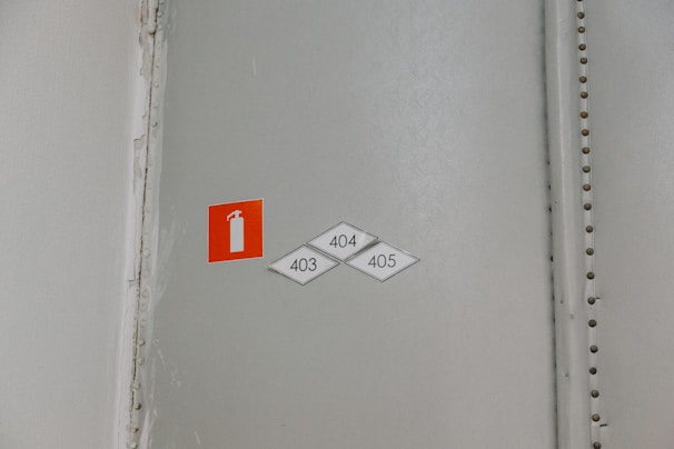 A wall with a small, rectangular red sign featuring a white fire extinguisher icon. Below the sign are three diamond-shaped labels with the numbers 403, 404, and 405. The wall is in a neutral color with a visible seam reinforced by metallic rivets.