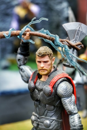 A highly detailed action figure of a superhero character wearing an armor with red accents and a cape. The figure holds an intricately designed hammer and has a focused expression. The background is blurred, highlighting the figure in the foreground.