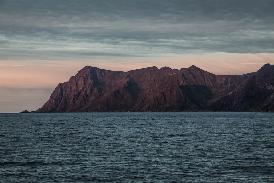 The image depicts a serene landscape with a large, dark brown mountain range stretching across the horizon. The mountains are partially obscured by clouds, adding depth and texture to the scene. The ocean is visible on the right side of the frame, its surface reflecting the colors of the sky above. The water appears calm, suggesting a peaceful atmosphere.