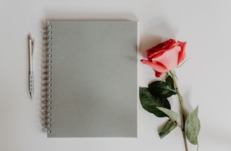 A spiral-bound notebook with a plain grey cover lies on a white surface, accompanied by a red rose with green leaves and a pen with a patterned grip.