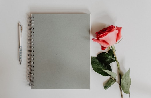 A spiral-bound notebook with a plain grey cover lies on a white surface, accompanied by a red rose with green leaves and a pen with a patterned grip.