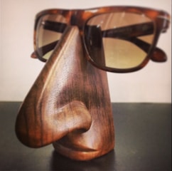 A wooden sculpture resembling a stylized human nose and lips serves as a stand for a pair of tortoiseshell sunglasses. The craftsmanship shows detailed wood grain and smooth contours, giving it a functional yet artistic appearance.