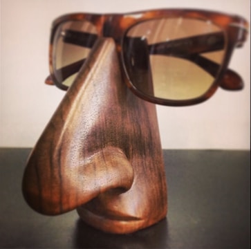 A wooden sculpture resembling a stylized human nose and lips serves as a stand for a pair of tortoiseshell sunglasses. The craftsmanship shows detailed wood grain and smooth contours, giving it a functional yet artistic appearance.