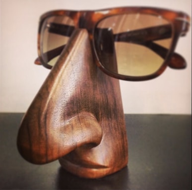 A wooden sculpture resembling a stylized human nose and lips serves as a stand for a pair of tortoiseshell sunglasses. The craftsmanship shows detailed wood grain and smooth contours, giving it a functional yet artistic appearance.