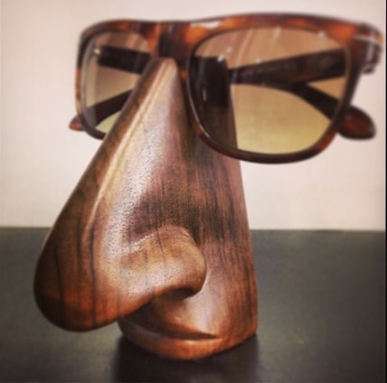 A wooden sculpture resembling a stylized human nose and lips serves as a stand for a pair of tortoiseshell sunglasses. The craftsmanship shows detailed wood grain and smooth contours, giving it a functional yet artistic appearance.