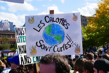 A crowd of people hold protest signs, one prominently featuring an illustration of the Earth surrounded by flames with text in French. The background consists of city buildings and trees with autumn foliage.