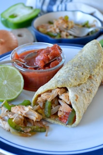 A rolled tortilla filled with shredded chicken, green bell peppers, and onions is placed on a plate. Beside it, there is a small bowl of chunky red salsa with a spoon. A half lime and fresh ingredients like a green bell pepper and onion are visible in the background, along with a bowl containing more of the chicken filling.