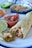 A rolled tortilla filled with shredded chicken, green bell peppers, and onions is placed on a plate. Beside it, there is a small bowl of chunky red salsa with a spoon. A half lime and fresh ingredients like a green bell pepper and onion are visible in the background, along with a bowl containing more of the chicken filling.