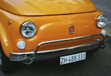 The close-up image features the front of a classic orange Fiat car. It focuses on the distinctive rounded headlights, chrome bumper, and the small Fiat emblem centrally placed on the metal grille. The car's registration plate is also visible at the bottom.