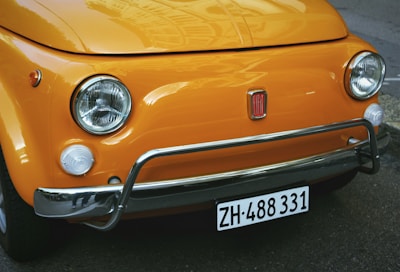 The close-up image features the front of a classic orange Fiat car. It focuses on the distinctive rounded headlights, chrome bumper, and the small Fiat emblem centrally placed on the metal grille. The car's registration plate is also visible at the bottom.