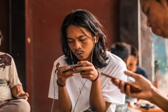 A person with long hair is intently using a smartphone, wearing earphones, and is surrounded by others in a warm, indoor environment. The focus is on the individual in the foreground, with others slightly blurred in the background. The ambiance suggests a casual or relaxed setting.
