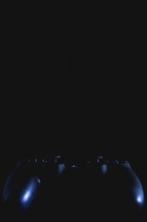 A dark and dimly lit image featuring the silhouette of a video game controller. The highlights reveal parts of the buttons and contours with subtle reflections.