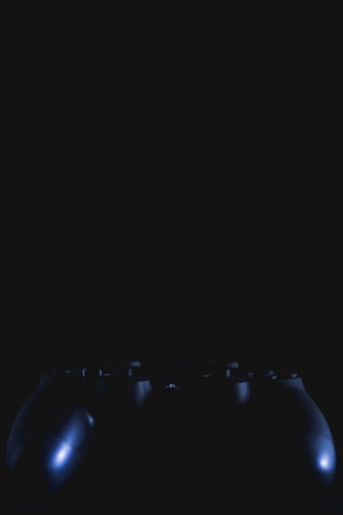 A dark and dimly lit image featuring the silhouette of a video game controller. The highlights reveal parts of the buttons and contours with subtle reflections.