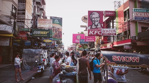 A bustling urban street scene featuring numerous storefronts and billboards advertising brands such as KFC and Subway. People are gathered around street vendors, engaging in activities like looking at phones and examining goods. The street is vibrant with signage in various colors, creating a lively, busy atmosphere.