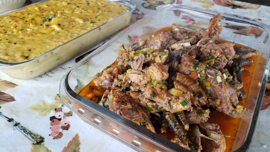 A table is set with two glass dishes containing food. One dish is filled with richly seasoned and cooked meat, garnished with chopped green herbs and sitting in a reddish-brown sauce. The second dish holds a creamy yellow mixture, possibly a casserole or pudding, with visible specks of herbs. Both dishes are placed on a patterned tablecloth with floral designs.