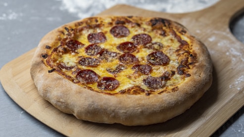 A freshly baked pepperoni pizza rests on a wooden pizza peel. The pizza features a golden-brown crust and is topped with melted cheese and evenly distributed pepperoni slices. The setting suggests a kitchen environment with a dusting of flour visible on the surface.