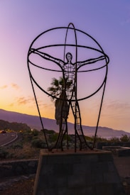 A metal sculpture in the shape of a human figure standing on a pedestal with arms outstretched within a large spherical frame. The background features a scenic view of a road, greenery, and distant mountains under a colorful, twilight sky with purple and orange hues.