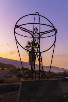 A metal sculpture in the shape of a human figure standing on a pedestal with arms outstretched within a large spherical frame. The background features a scenic view of a road, greenery, and distant mountains under a colorful, twilight sky with purple and orange hues.