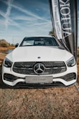 A white Mercedes-Benz GLC Coupé is prominently displayed on a grassy field. The car's front grille features the iconic Mercedes emblem. A large vertical banner is positioned beside the vehicle, partially obscuring the view.