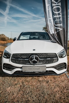 A white Mercedes-Benz GLC Coupé is prominently displayed on a grassy field. The car's front grille features the iconic Mercedes emblem. A large vertical banner is positioned beside the vehicle, partially obscuring the view.