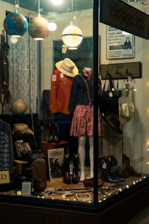 A shop window display with mannequins wearing vintage clothing, including a red jacket and a floral skirt. Various items such as hats, boots, and globes are arranged alongside decorative string lights. The setting is cozy and somewhat nostalgic, evoking a sense of old-fashioned charm.