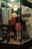 A shop window display with mannequins wearing vintage clothing, including a red jacket and a floral skirt. Various items such as hats, boots, and globes are arranged alongside decorative string lights. The setting is cozy and somewhat nostalgic, evoking a sense of old-fashioned charm.