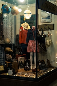 A shop window display with mannequins wearing vintage clothing, including a red jacket and a floral skirt. Various items such as hats, boots, and globes are arranged alongside decorative string lights. The setting is cozy and somewhat nostalgic, evoking a sense of old-fashioned charm.