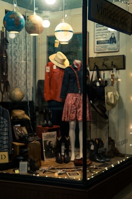 A shop window display with mannequins wearing vintage clothing, including a red jacket and a floral skirt. Various items such as hats, boots, and globes are arranged alongside decorative string lights. The setting is cozy and somewhat nostalgic, evoking a sense of old-fashioned charm.