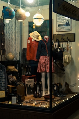 A shop window display with mannequins wearing vintage clothing, including a red jacket and a floral skirt. Various items such as hats, boots, and globes are arranged alongside decorative string lights. The setting is cozy and somewhat nostalgic, evoking a sense of old-fashioned charm.