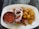A white plate carries a serving of grilled skewered meat with sliced onions, bell peppers, and roasted potatoes. A small metal bowl contains a spicy red sauce, while a powder condiment is also present on the plate.
