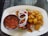 A white plate carries a serving of grilled skewered meat with sliced onions, bell peppers, and roasted potatoes. A small metal bowl contains a spicy red sauce, while a powder condiment is also present on the plate.