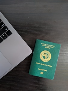 A green Nigerian passport is placed on a dark wooden table beside the corner of a laptop. The passport features gold text and a circular emblem representing the Economic Community of West African States (ECOWAS) at the center.
