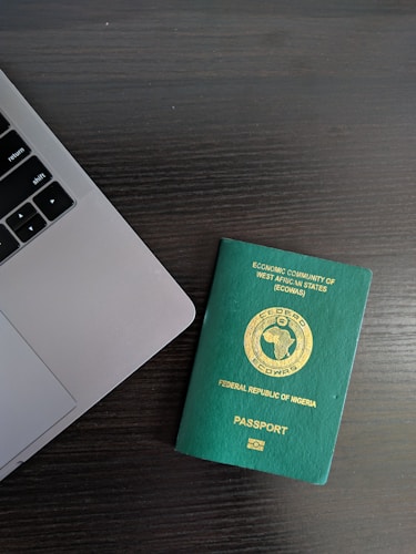 A green Nigerian passport is placed on a dark wooden table beside the corner of a laptop. The passport features gold text and a circular emblem representing the Economic Community of West African States (ECOWAS) at the center.