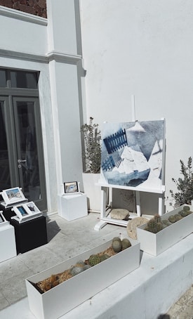 An outdoor art display features a large canvas on an easel showing a painting with geometric shapes and shades of blue and white. Several smaller framed artworks are arranged on pedestals nearby. Potted cacti and small plants are placed in rectangular planters, adding a touch of greenery against the minimalistic architecture of white walls with some modernist design elements.