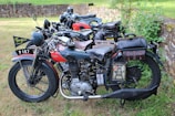 A collection of vintage motorcycles parked in a grassy outdoor area. These motorcycles appear well-maintained, featuring leather saddlebags and classic design elements such as round headlights and large tires. The setting includes a stone wall and foliage, suggesting a rural or historical environment.