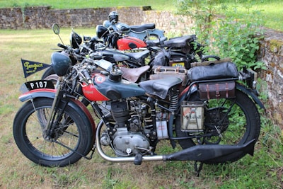 A collection of vintage motorcycles parked in a grassy outdoor area. These motorcycles appear well-maintained, featuring leather saddlebags and classic design elements such as round headlights and large tires. The setting includes a stone wall and foliage, suggesting a rural or historical environment.