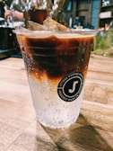 A plastic cup filled with a layered iced coffee drink sits on a wooden table. The drink has a gradient from a dark brown coffee at the top to sparkling water at the bottom. Ice cubes are visible at the top, and bubbles can be seen in the clear section of the drink. The cup features a logo with the text 'Jitterz Coffee Roasters Proudly Made in Texas.' The background has blurred elements suggesting a cozy café setting.
