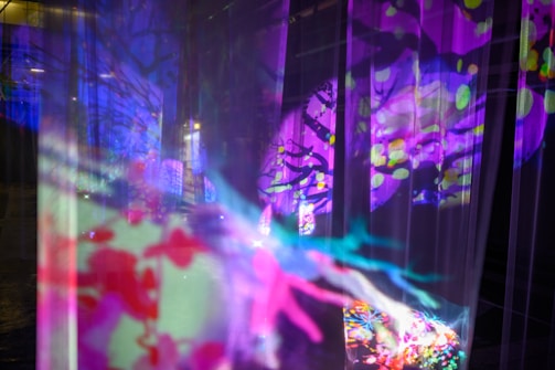 Vibrant colors and abstract patterns create a dynamic and immersive visual experience. The image features overlapping layers of translucent fabric or screens, onto which bright, saturated colors like purple, pink, green, and blue are projected. Organic shapes and abstract designs evoke a sense of movement and fluidity, possibly suggesting elements of nature or fantasy.