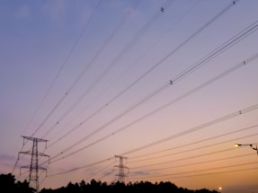 A network of power lines stretches across the sky, connected by tall metal pylons. The sky is transitioning from shades of purple to orange, indicating either sunrise or sunset. Trees are silhouetted against the horizon, and a streetlamp is visible in the lower right corner, casting a warm glow.