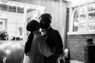 A couple is seen kissing in a cozy setting with soft lighting. The image has a warm and intimate indoor ambiance with string lights decorating the background. The people are dressed warmly, suggesting a cool or winter evening.