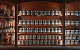 A wooden shelf is stocked with numerous jars and packages of food products. The shelves are neatly organized with rows of jars featuring labels and metallic lids. To the left, various packaged goods such as pastas and spices are visible. The lighting creates a warm, inviting atmosphere, highlighting the rich tones of the wood.