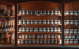 A wooden shelf is stocked with numerous jars and packages of food products. The shelves are neatly organized with rows of jars featuring labels and metallic lids. To the left, various packaged goods such as pastas and spices are visible. The lighting creates a warm, inviting atmosphere, highlighting the rich tones of the wood.