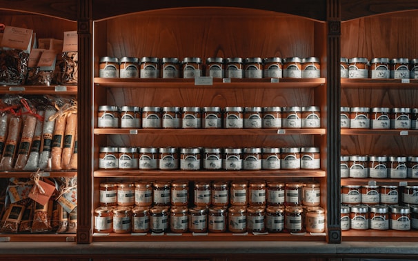 A wooden shelf is stocked with numerous jars and packages of food products. The shelves are neatly organized with rows of jars featuring labels and metallic lids. To the left, various packaged goods such as pastas and spices are visible. The lighting creates a warm, inviting atmosphere, highlighting the rich tones of the wood.
