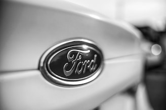 Close-up view of a Ford logo on the front of a vehicle, with a focus on the emblem and a blurred background.