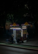 A street food cart is illuminated on a dimly lit sidewalk at night. The cart has colorful pictures of menu items and bright lights highlighting its structure. Two people are near the cart, possibly a vendor inside and a customer outside.