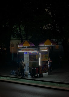 A street food cart is illuminated on a dimly lit sidewalk at night. The cart has colorful pictures of menu items and bright lights highlighting its structure. Two people are near the cart, possibly a vendor inside and a customer outside.