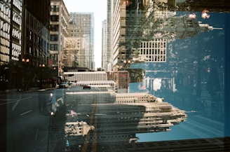 A visually intriguing double exposure photograph featuring urban buildings with various architectural styles. The image is a composite of city structures overlapping, reflecting a dynamic cityscape. Glass facades and streetlights are visible, creating a mix of reflections and clear depictions of the buildings.
