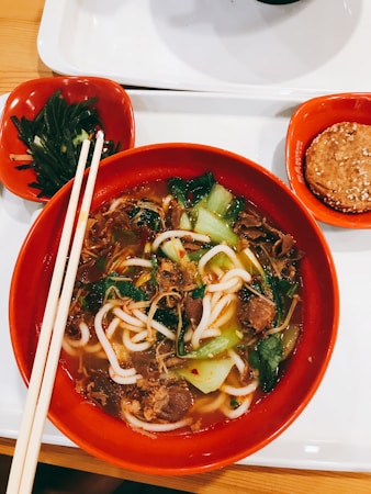 A bowl of noodle soup with a variety of ingredients including green vegetables, sliced meat, and noodles, served in a red bowl with chopsticks resting on the side. Accompanying the main dish are side servings of seaweed salad and a sesame cookie, each in its own small red dish, all placed on a white tray.