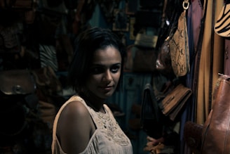 A woman with short dark hair gazes at the camera in a dimly lit room filled with leather bags hanging on the walls. The lighting casts shadows that create a moody atmosphere. She is wearing a cream-colored sleeveless top.