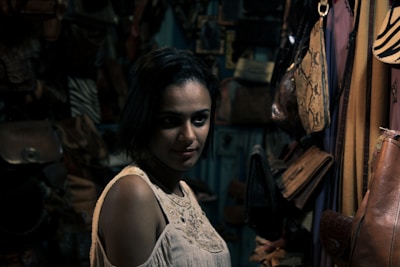 A woman with short dark hair gazes at the camera in a dimly lit room filled with leather bags hanging on the walls. The lighting casts shadows that create a moody atmosphere. She is wearing a cream-colored sleeveless top.